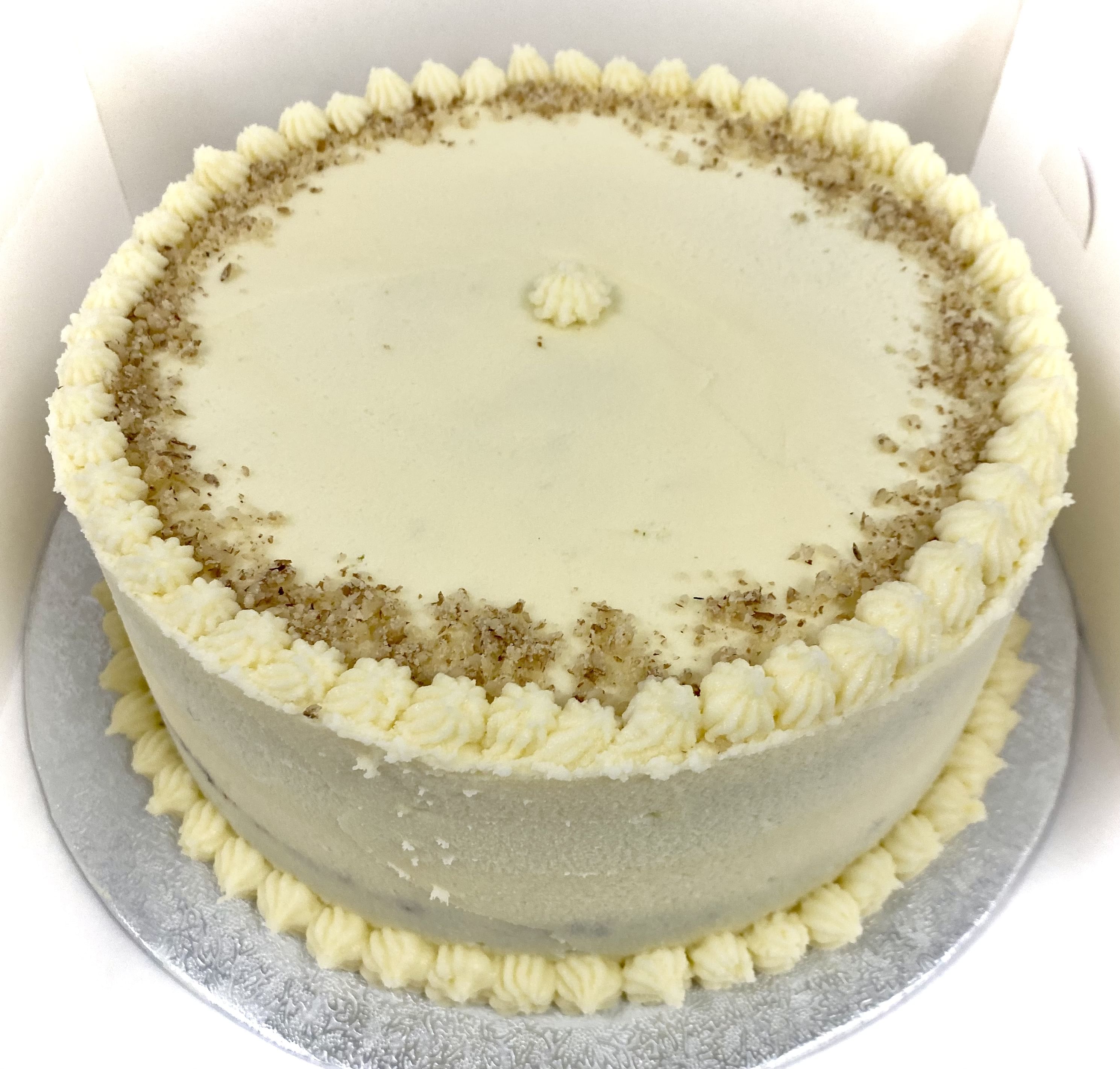 A frosted round cake with a light beige color, displayed on a silver-colored board. It has decorative piped icing along the top edge and a circle of finely ground nuts near the edges, with a dollop of icing in the center. This award-winning carrot cake is truly delightful.